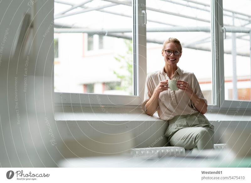 Porträt einer lächelnden jungen Geschäftsfrau mit Tasse Kaffee am Fenster sitzend Job Berufe Berufstätigkeit Beschäftigung Jobs geschäftlich Geschäftsleben