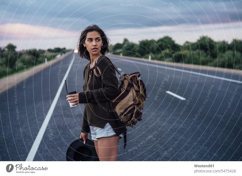 Porträt einer jungen Frau beim Trampen mit Rucksack und Getränk auf der Fahrbahn stehend Rucksäcke weiblich Frauen trampen Anhalter per Autostopp fahren