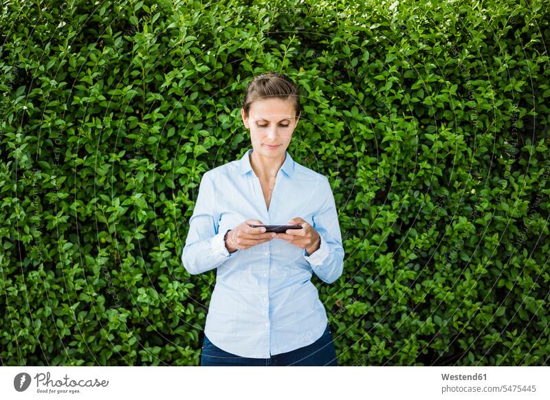 Frau steht mit Mobiltelefon an einer Hecke stehen stehend Hecken Handy Handies Handys Mobiltelefone weiblich Frauen Telefon telefonieren Kommunikation