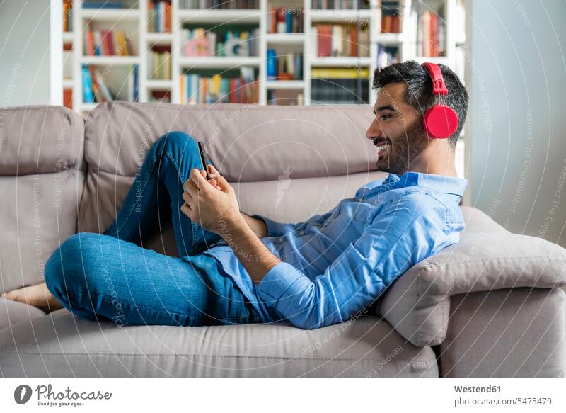 Lächelnder junger Mann, der zu Hause auf der Couch liegt und ein Smartphone und drahtlose Kopfhörer benutzt Leute Menschen People Person Personen Europäisch