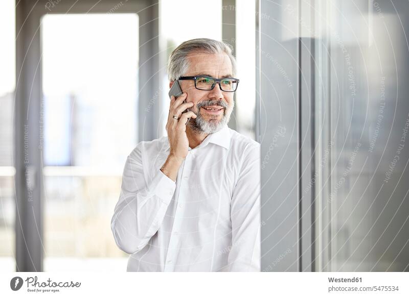 Lächelnder Geschäftsmann spricht am Fenster mit dem Handy stehen stehend steht telefonieren anrufen Anruf telephonieren Mobiltelefon Handies Handys