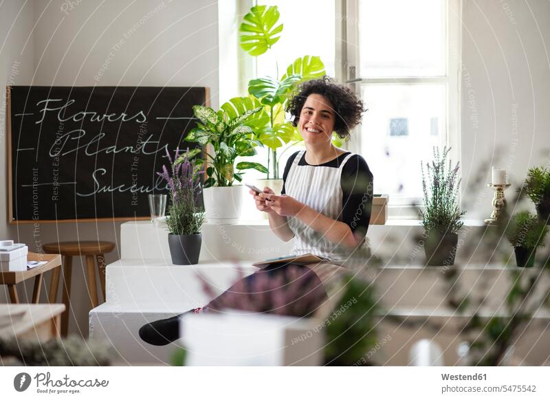 Porträt einer lächelnden jungen Frau, die ein Mobiltelefon in einem kleinen Laden mit Pflanzen benutzt Job Berufe Berufstätigkeit Beschäftigung Jobs Leute