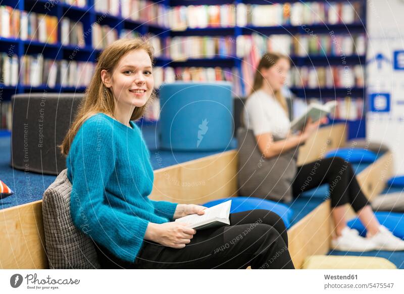 Porträt eines lächelnden Teenager-Mädchens, das in einer öffentlichen Bibliothek sitzt und ein Buch liest Teenagerin junges Mädchen Teenagerinnen weiblich