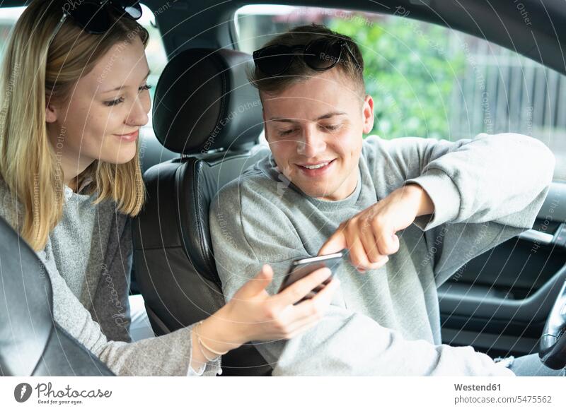 Lächelndes junges Paar im Auto schaut auf Handy Leute Menschen People Person Personen Kraftfahrer Informationen KFZ Verkehrsmittel Automobil Autos PKW PKWs
