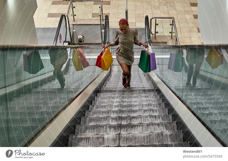 Glückliche schwarze Frau mit lustiger Frisur, die bunte Einkaufstaschen in der Hand hält und auf den Rolltreppen läuft Taschen Tuete Tueten Tüten Einkaufstüten