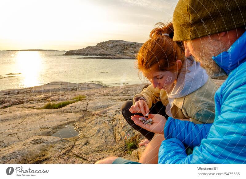 Schweden, Grafschaft Vastra Gotaland, Grebbestad, Mann und Jugendliche sammeln bei Sonnenuntergang Kieselsteine am felsigen Ufer des Naturreservats Tjurpannans