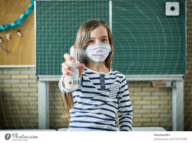 Porträt eines maskierten Mädchens, das im Klassenzimmer Desinfektionsmittel sprüht Ausbildung Schueler Schulkinder Schüler Schultafel Schultafeln Gesund