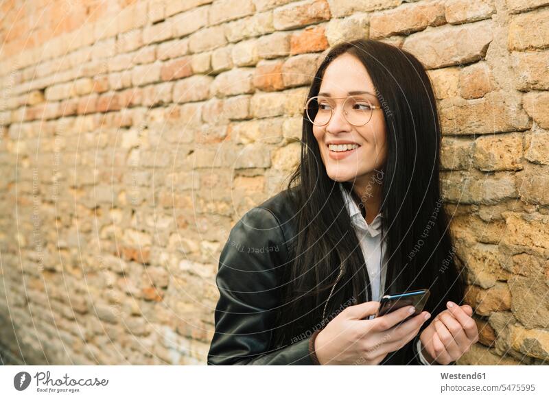 Lächelnde junge Frau mit Handy an Ziegelmauer, die sich umsieht umherblicken umschauen umsehen Mobiltelefon Handies Handys Mobiltelefone weiblich Frauen lächeln