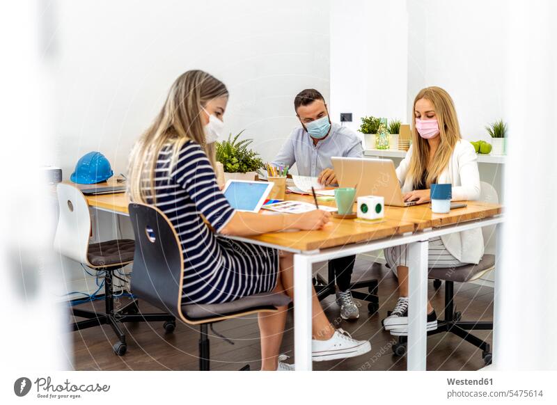 Geschäftsleute, die im Büro am Schreibtisch sitzen und eine Gesichtsmaske tragen Farbaufnahme Farbe Farbfoto Farbphoto Innenaufnahme Innenaufnahmen innen