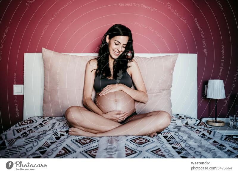 Glückliche schwangere Frau schaut auf den Bauch, während sie zu Hause im Schlafzimmer sitzt Farbaufnahme Farbe Farbfoto Farbphoto Innenaufnahme Innenaufnahmen