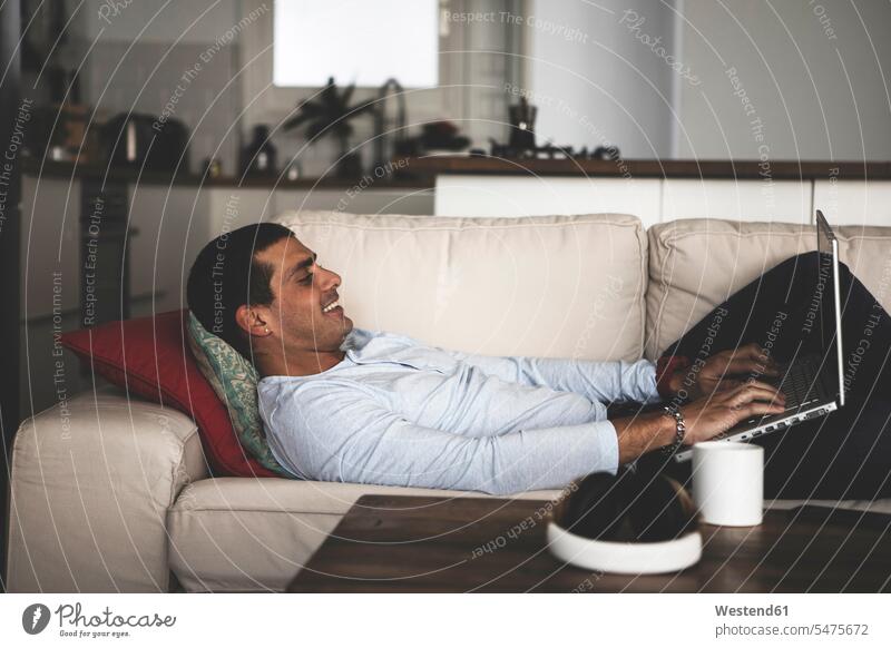 Lächelnder junger Mann auf dem Sofa zu Hause liegend mit Laptop Männer männlich lächeln liegt Couches Liege Sofas Notebook Laptops Notebooks Zuhause daheim