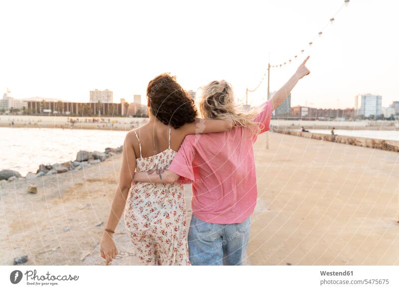 Rückansicht von zwei jungen Frauen auf der Uferpromenade bei Sonnenuntergang Freunde Kameradschaft Touristen T-Shirts gehend geht Arm umlegen Umarmung