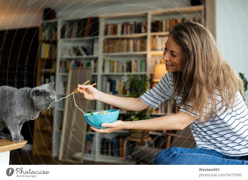 Lächelnde junge Frau füttert Katze mit Spaghetti, während sie zu Hause sitzt Farbaufnahme Farbe Farbfoto Farbphoto Spanien Freizeitkleidung Freizeitbekleidung