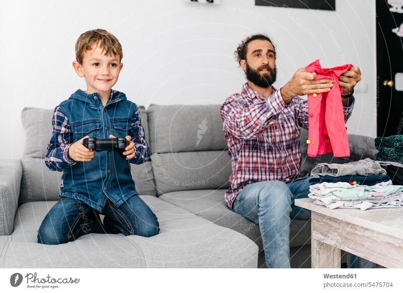 Porträt eines lächelnden kleinen Jungen, der ein Computerspiel spielt, während sein Vater Wäsche faltet Väter Papa Vati Familienvater Papi Buben Knabe Knaben