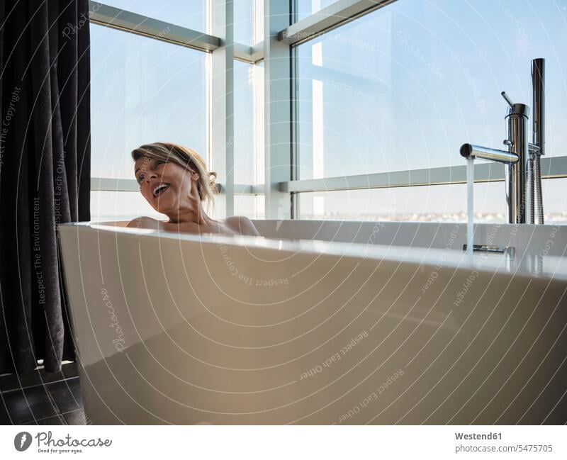 Fröhliche blonde ältere Frau badet in Badewanne gegen Fenster in Luxus-Hotelzimmer Farbaufnahme Farbe Farbfoto Farbphoto Innenaufnahme Innenaufnahmen innen