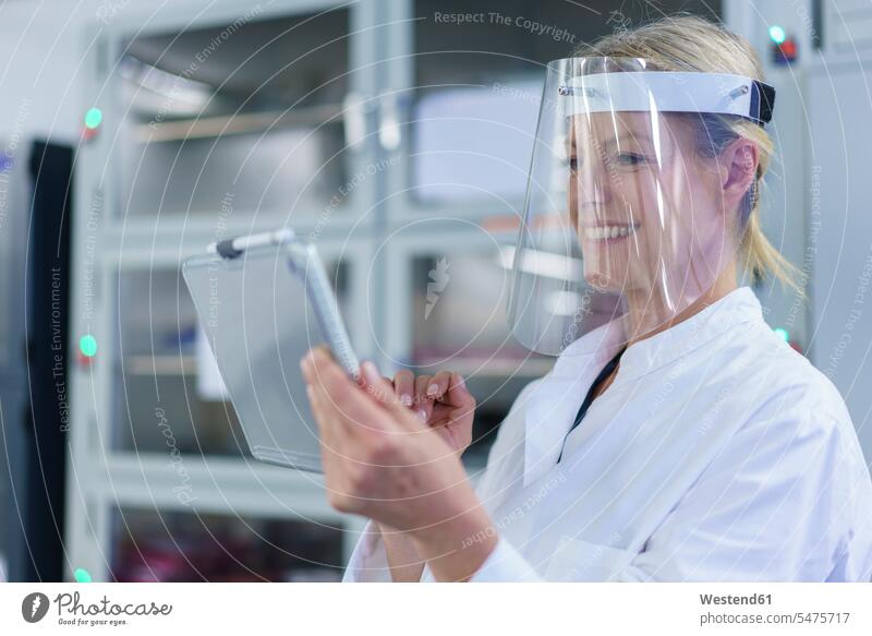 Lächelnde reife Wissenschaftlerin mit Gesichtsschutz bei der Verwendung eines digitalen Tabletts im Labor Farbaufnahme Farbe Farbfoto Farbphoto Innenaufnahme