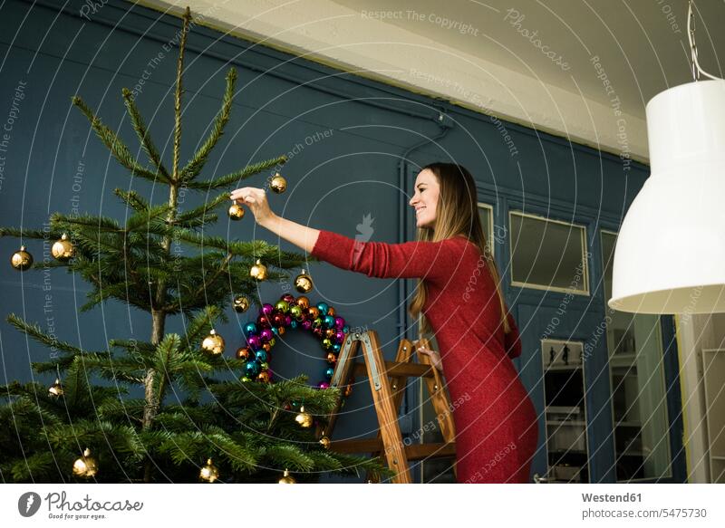Lächelnde Frau steht auf Leiter und schmückt Weihnachtsbaum stehen stehend Leitern Christbaum Weihnachtsbäume Weihnachtsbaeume Christbäume Christbaeume weiblich
