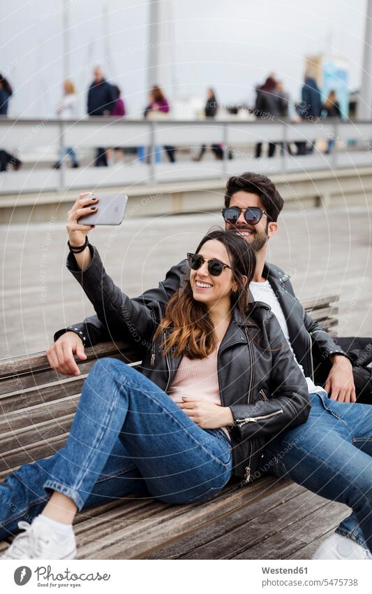 Spanien, Barcelona, glückliches junges Paar, das sich auf einer Bank ausruht und ein Selfie macht Sitzbänke Bänke Sitzbank ausruhen Rast Erholung erholen
