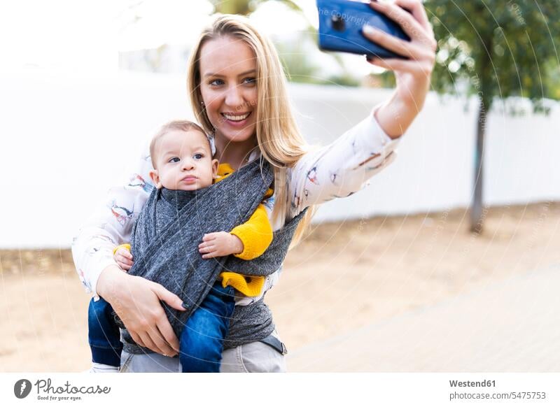 Glückliche Mutter mit Baby in einem Tragetuch macht ein Selfie Telekommunikation telefonieren Handies Handys Mobiltelefon Mobiltelefone freuen Frohsinn