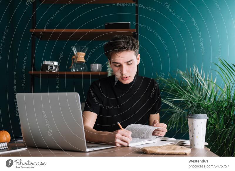 Männlicher Student schreibt in Buch, während er mit dem Laptop am Tisch sitzt und Hausaufgaben macht Farbaufnahme Farbe Farbfoto Farbphoto Innenaufnahme