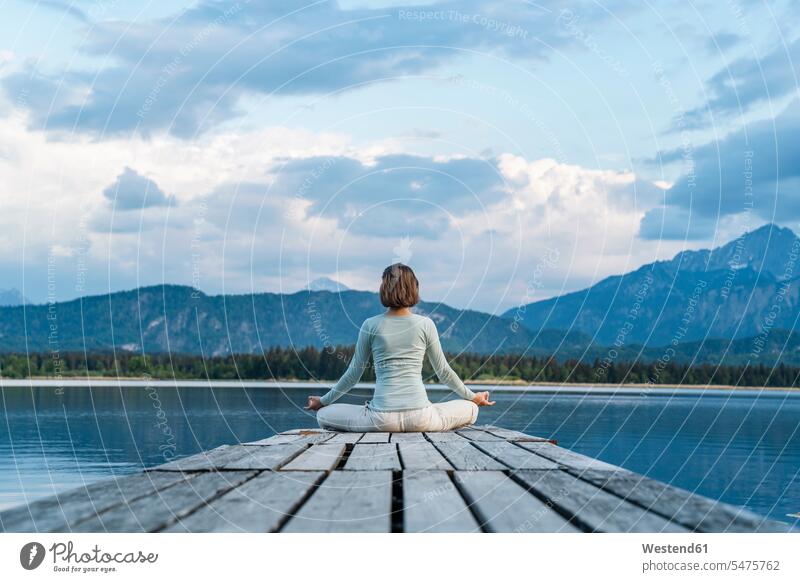 Mittelgroße erwachsene Frau meditiert, während sie auf einem Steg über dem See vor bewölktem Himmel sitzt Farbaufnahme Farbe Farbfoto Farbphoto Deutschland