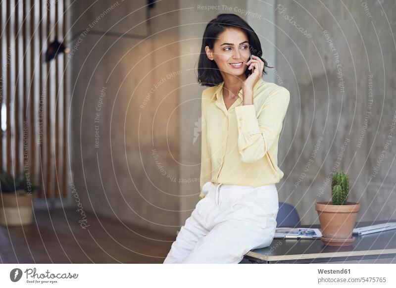 Lächelnde Unternehmerin spricht über Mobiltelefon, während sie im Büro am Schreibtisch sitzt Farbaufnahme Farbe Farbfoto Farbphoto Deutschland Innenaufnahme