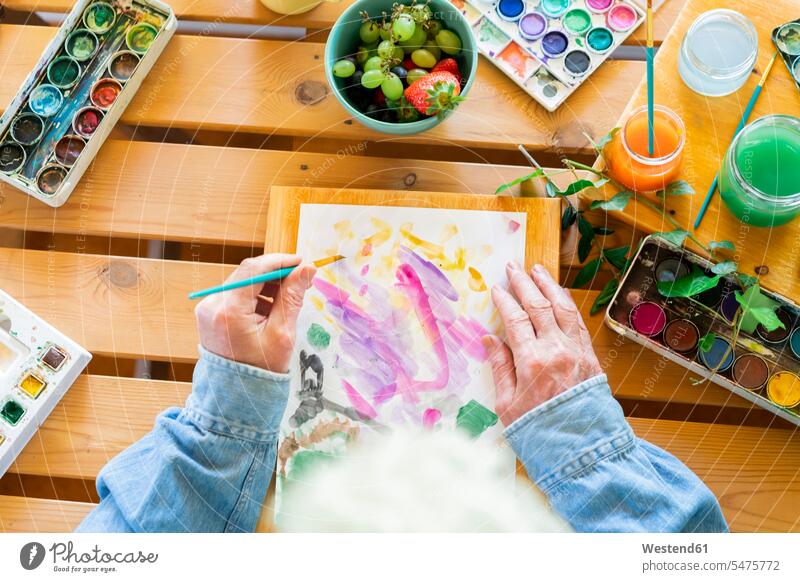 Älterer Mann malt auf Papier am Holztisch Farbaufnahme Farbe Farbfoto Farbphoto Innenaufnahme Innenaufnahmen innen drinnen Tag Tageslichtaufnahme