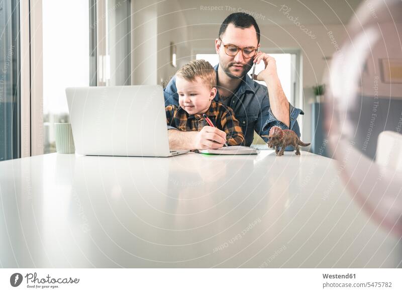 Beschäftigter Vater arbeitet am Tisch im Home-Office mit Sohn sitzt auf dem Schoß sitzen sitzend Söhne arbeiten Arbeit Papas Väter Vati Vatis Papis