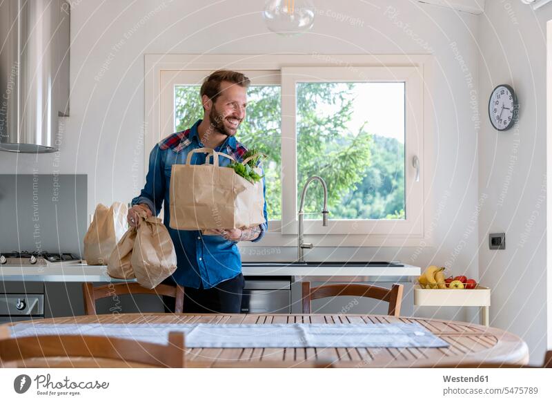 Lächelnder, gutaussehender Mann hält Einkaufstasche in der Hand, während er zu Hause in der Küche wegschaut Farbaufnahme Farbe Farbfoto Farbphoto Innenaufnahme