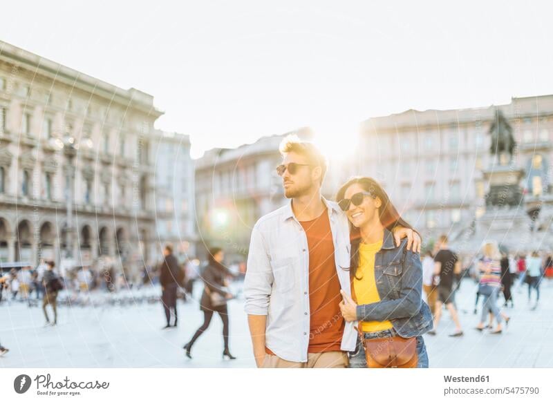 Glückliches junges Paar auf einem Platz in der Stadt bei Sonnenuntergang, Mailand, Italien Touristen Brillen Sonnenbrillen Arm umlegen Umarmung Umarmungen