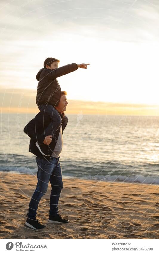 Vater trägt seinen Sohn bei Sonnenuntergang huckepack am Strand und zeigt mit dem Finger tragen transportieren Sonnenuntergänge deuten Huckepack Papas Väter