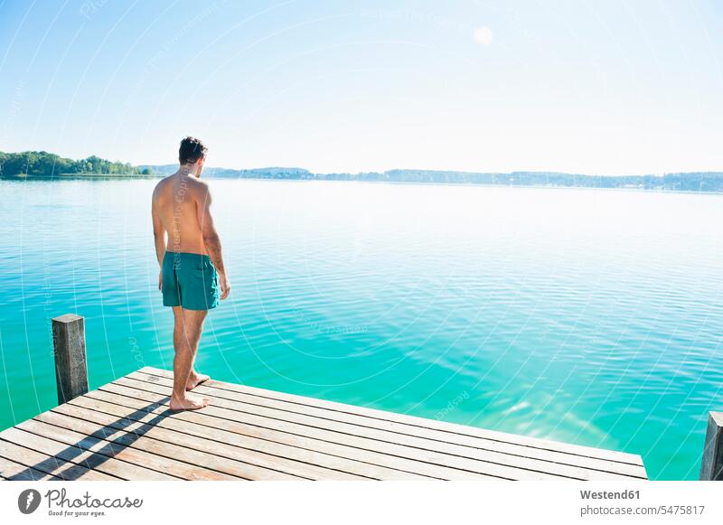Rückenansicht eines jungen Mannes in Badeshorts, der auf einem Steg steht und auf den See schaut entspannen relaxen Jahreszeiten sommerlich Sommerzeit stehend