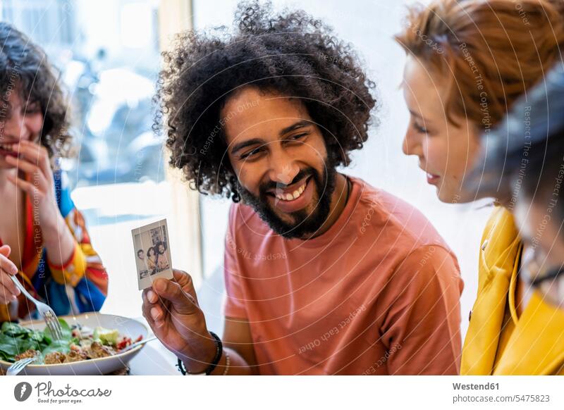 Glücklicher Mann zeigt Freunden beim Mittagessen in einem Restaurant ein Sofortbild Bilder Bildnis Fotos Polaroid Polaroids Sofortbilder Tische sitzend sitzt
