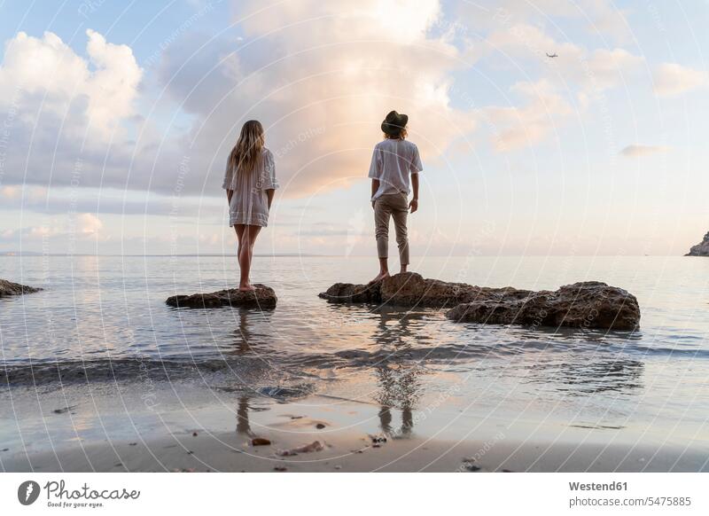 Rückenansicht eines jungen Paares, das auf Felsen vor dem Meer steht und ein Flugzeug beobachtet, Ibiza, Balearen, Spanien Flieger Flugzeuge entspannen relaxen