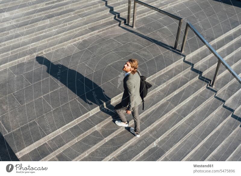 Männlicher Unternehmer mit einer Tasche, der sich auf der Treppe nach oben bewegt, während er an einem sonnigen Tag in der Innenstadt wegschaut Farbaufnahme