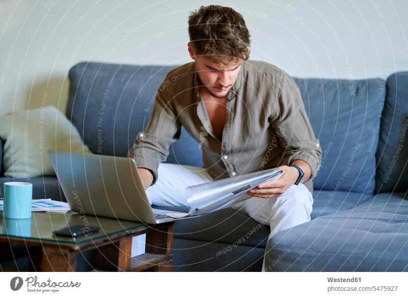 Junger männlicher Freiberufler liest Dokumente, während er zu Hause auf einem Sofa mit Laptop sitzt Farbaufnahme Farbe Farbfoto Farbphoto Innenaufnahme