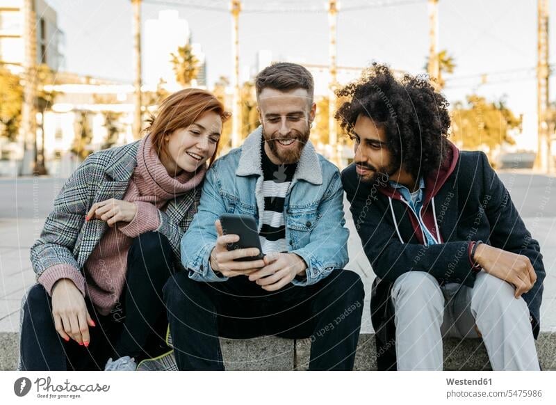 Drei glückliche Freunde sitzen im Freien und schauen auf ihr Handy Glück glücklich sein glücklichsein sitzend sitzt ansehen Mobiltelefon Handies Handys