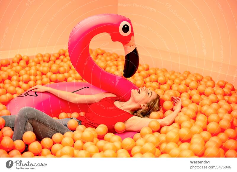 Fröhliche junge Frau mit aufblasbarem Flamingo in orangefarbener Ballgrube liegend Farbaufnahme Farbe Farbfoto Farbphoto Freizeitbeschäftigung Muße Zeit