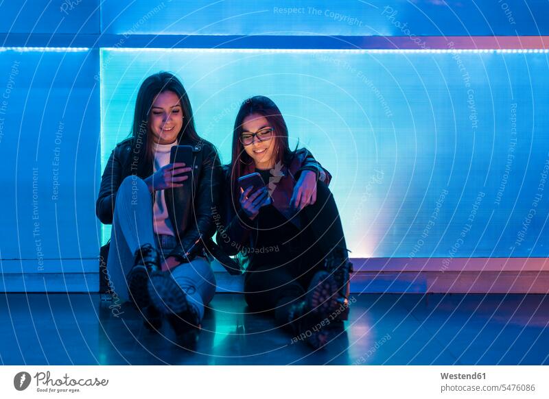 Porträt von zwei lächelnden Teenager-Mädchen, die vor einer blauen Glasscheibe sitzen und auf Smartphones schauen Leute Menschen People Person Personen