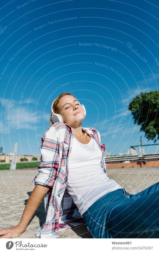 Frau mit geschlossenen Augen, die bei Sonnenschein über Kopfhörer Musik hört und auf dem Fußweg sitzt Farbaufnahme Farbe Farbfoto Farbphoto Außenaufnahme außen