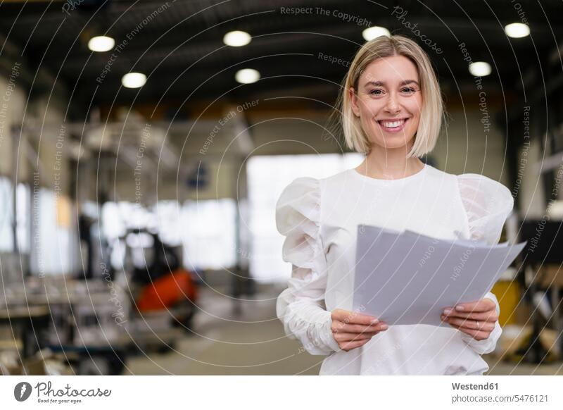 Porträt einer lächelnden jungen Frau, die in einer Fabrik Papiere in der Hand hält Leute Menschen People Person Personen Europäisch Kaukasier kaukasisch 1 Ein
