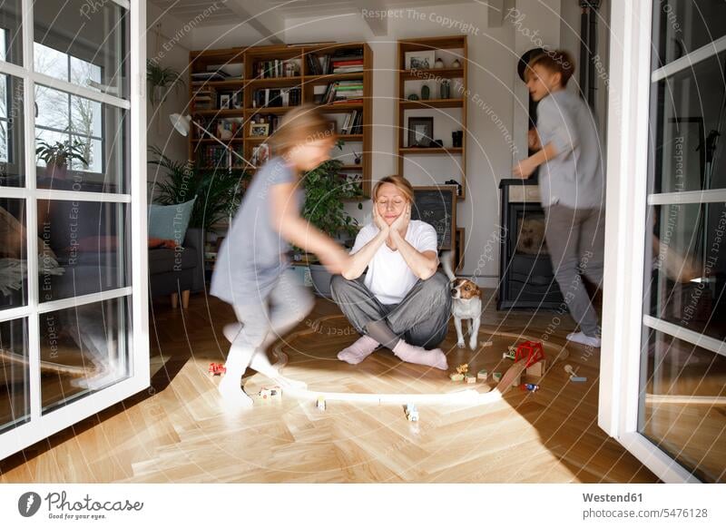 Gestresste Mutter, die in der Mitte des Spielzeugs sitzt, während die Kinder um sie herumlaufen rennen sitzend Kreise daheim zu Hause abgespannt geöffnet