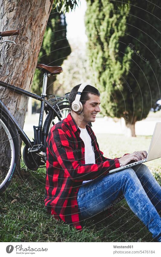 Junger Mann sitzt in einem Park mit Kopfhörern und mit Laptop neben dem Fahrrad Männer männlich Notebook Laptops Notebooks Bikes Fahrräder Räder Rad Parkanlagen