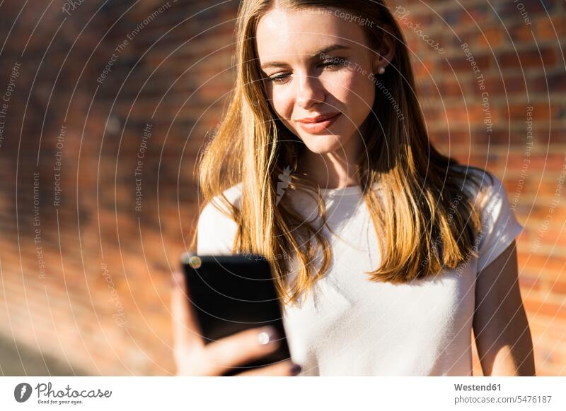 Junge Frau vor einer Backsteinmauer, mit Smartphone stehen stehend steht iPhone Smartphones benutzen benützen Nachricht Mitteilung Botschaft junge Frau