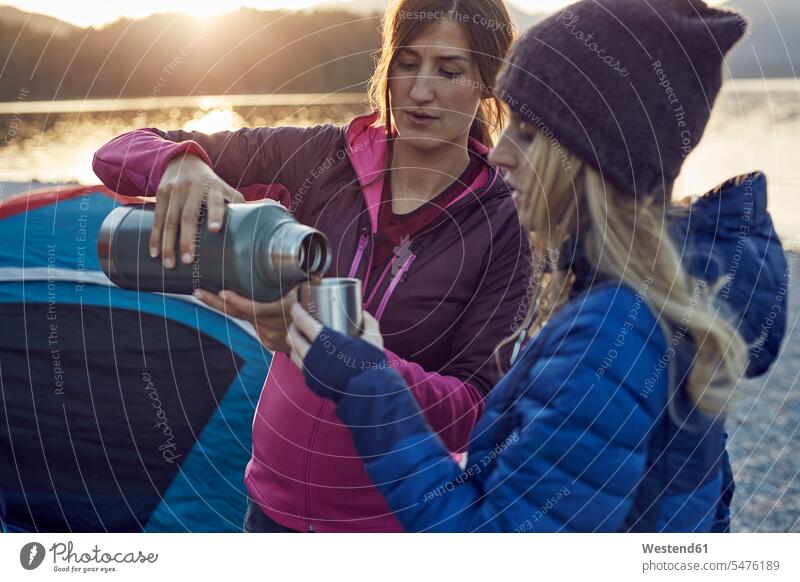 Zwei Frauen mit heißen Getränken zelten am Seeufer Freundinnen Zelt Zelte weiblich Heißgetränk Heissgetraenk Heissgetraenke Heißgetränke Camping Campen Freunde