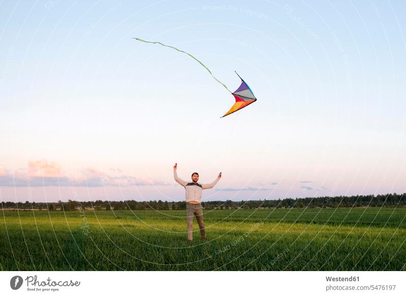 Mittelgroßer erwachsener Mann fliegt Drachen, während er bei Sonnenuntergang auf einer Graslandschaft gegen den Himmel steht Farbaufnahme Farbe Farbfoto
