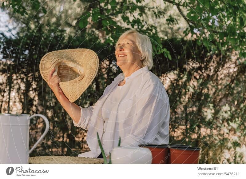 Fröhliche ältere Frau mit Hut in der Hand schaut auf, während sie im Hof an einem Baum sitzt Farbaufnahme Farbe Farbfoto Farbphoto Spanien Freizeitbeschäftigung