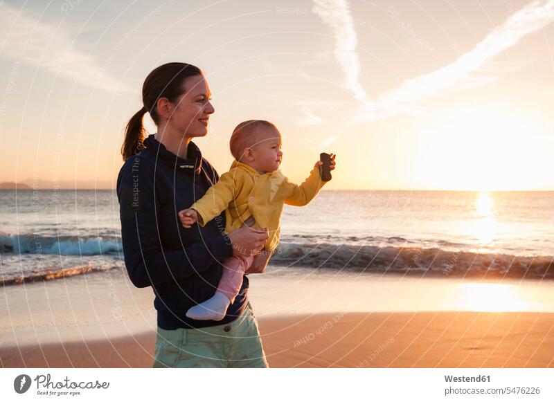 Mutter hält kleine Tochter bei Sonnenuntergang am Strand Beach Straende Strände Beaches Sonnenuntergänge Mami Mutti Mütter Mama Töchter halten Baby Babies Babys