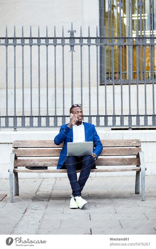 Junger Geschäftsmann in blauer Anzugjacke sitzt auf Bank und benutzt Smartphone Leute Menschen People Person Personen Afrikanisch Afrikanische Abstammung