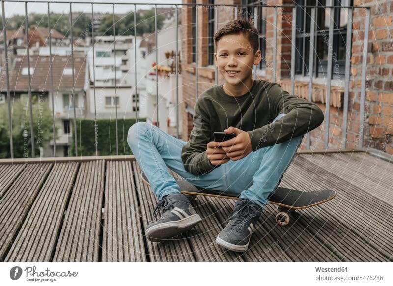 Lächelnder Junge benutzt Smartphone, während er auf einem Skateboard gegen die Reling sitzt Farbaufnahme Farbe Farbfoto Farbphoto Deutschland